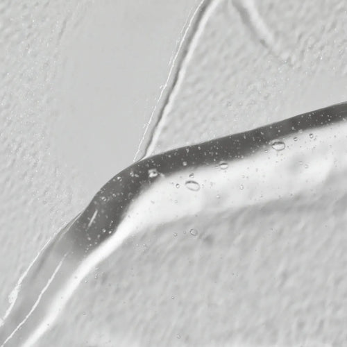 Grande plano do Solawaves LightBoost Wand Activating Serum, um gel transparente infundido com ácido hialurónico. Apresenta bolhas de ar contra um fundo branco texturado, realçando a superfície lisa e brilhante em contraste com o fundo rugoso.